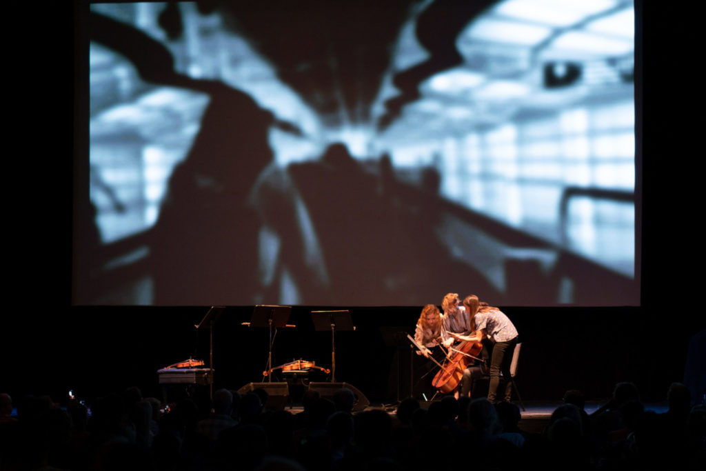 Holly Workman, violin, Pperforming Pamela Z Attention on one cello with three others in front of a large video projection
