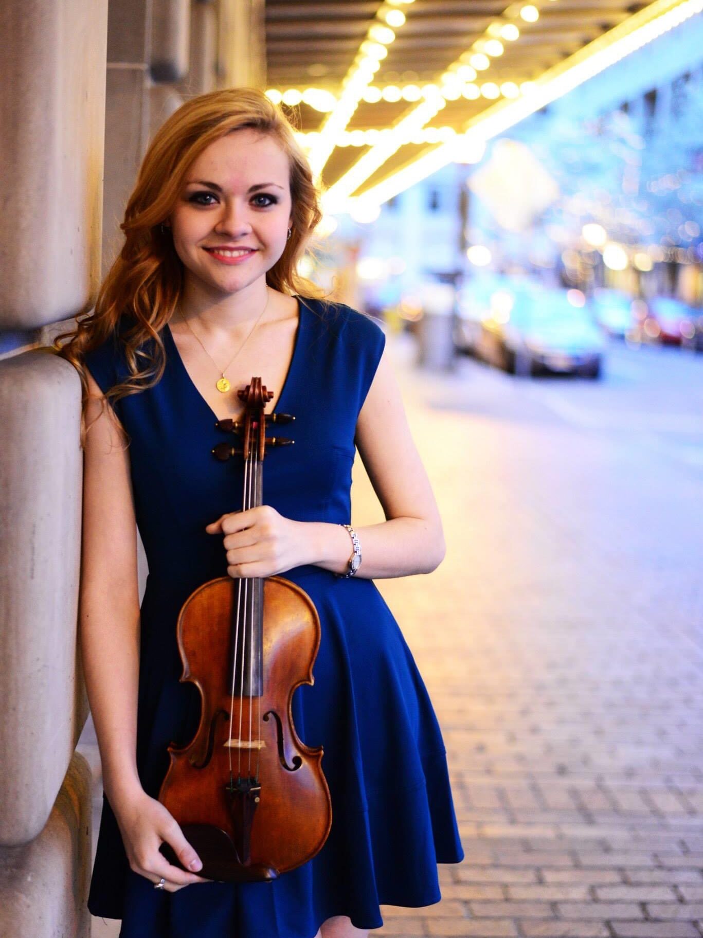 Holly Workman headshot, holding a violin, outside under theater lights
